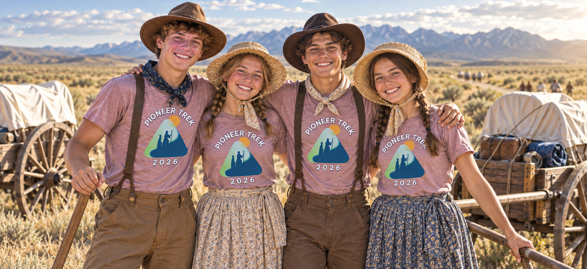 Four smiling teenagers wearing matching custom Pioneer Trek 2026 t-shirts and pioneer-style clothing stand arm-in-arm on a Wyoming prairie with handcarts and mountains in the background.